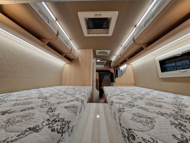 Wnętrze kampera z dwiema oddzielnymi łóżkami, drewnianymi szafkami i oświetleniem LED