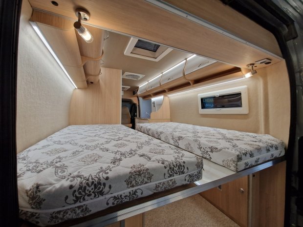 Wnętrze kampera z dwiema materacami, drewnianymi szafkami i oświetleniem LED