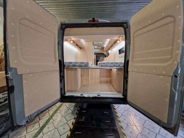 Otwarty tył kampera z dwoma łóżkami, jasnymi drewnianymi szafkami i przestrzenią bagażową