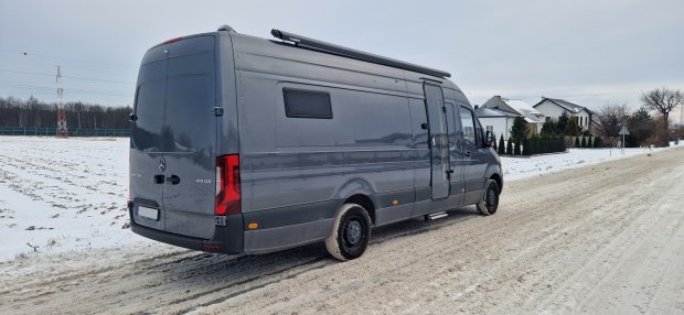 Szary Mercedes Sprinter zaparkowany na zaśnieżonej drodze przy domach.