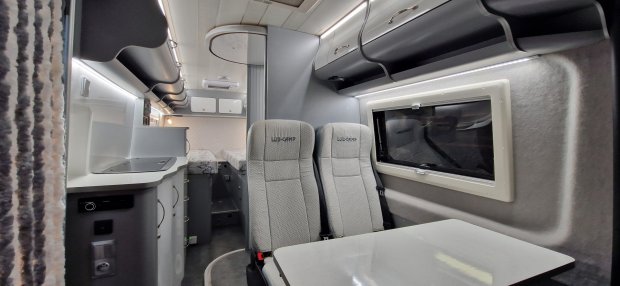 Wnętrze kampera z dwiema fotelami, stolikiem, kuchenką i tylnym łóżkiem