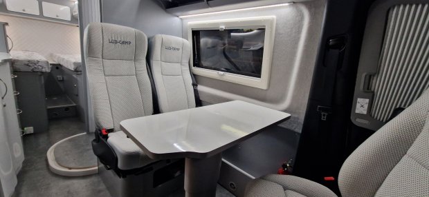 Wnętrze kampera: stół przy dwóch jasnych fotelach, okno i łóżko w tle