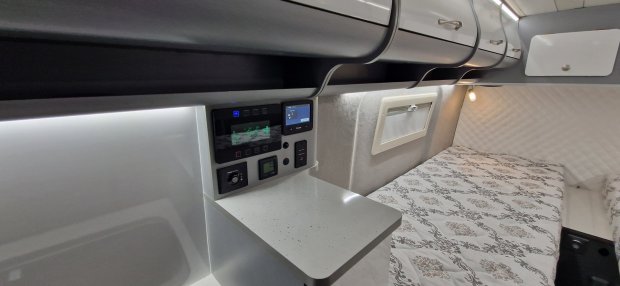 Wnętrze kampera: łóżko z wzorzystą pościelą, półki, panel sterowania i lampka.