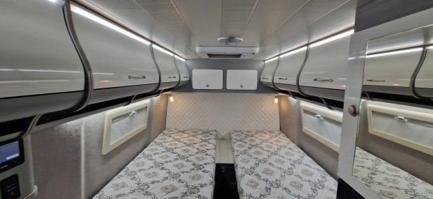 Wnętrze kampera z dwiema pojedynczymi kojami, górnymi szafkami i pikowanym zagłówkiem