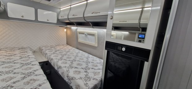 Wnętrze kampera z dwiema łóżkami, szafkami nad nimi i małym oknem