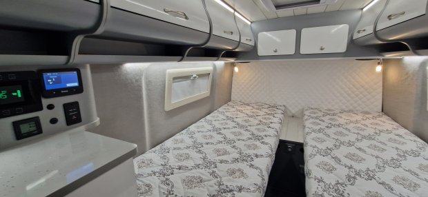 Wnętrze kampera: dwa pojedyncze łóżka, szafki nad nimi, lampki i małe okno.