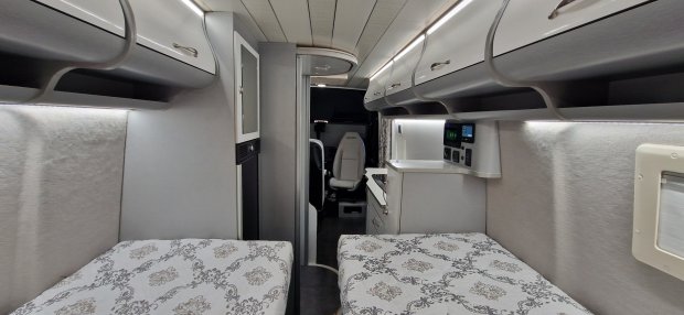 Wnętrze kampera z dwiema łóżkami, górnymi schowkami i niewielką kuchnią.