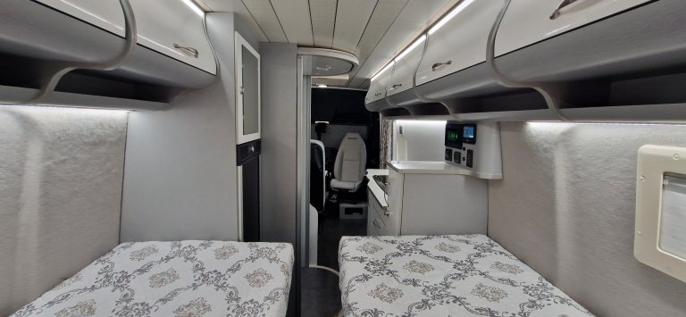 Wnętrze kampera z dwiema łóżkami, górnymi schowkami i niewielką kuchnią.