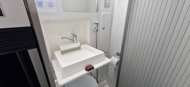 Mała biała umywalka z chromowaną baterią, prysznicem ręcznym i rolką papieru toaletowego.