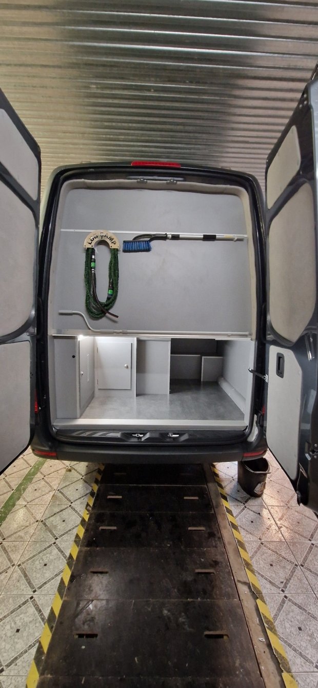 Otwarty tył furgonu z szarymi schowkami, szczotką i zwiniętym zielonym sznurem na ścianie.