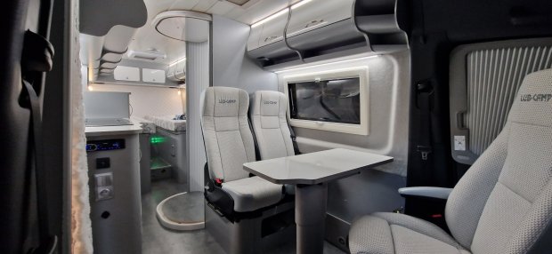 Wnętrze kampera: szare obrotowe fotele, stolik, schowki i łóżko w tle.