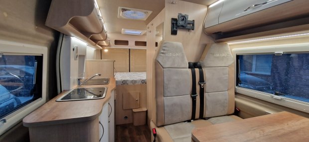 Wnętrze kampera: aneks kuchenny, dwa fotele z pasami i łóżko w tle.