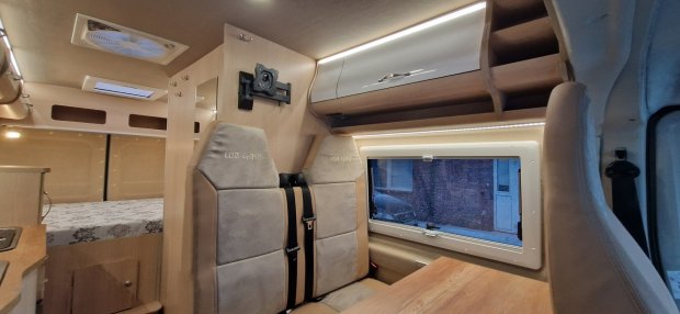 Wnętrze kampera: beżowe siedzenia z pasami, okno, szafki i część łóżka.