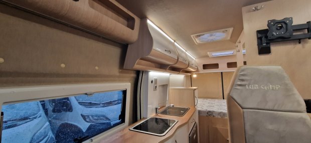 Wnętrze kampera z aneksem kuchennym, szafkami górnymi, zlewem i łóżkiem w tle.