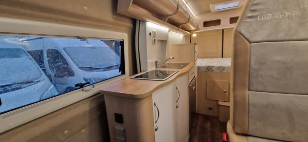 Wnętrze kampera: aneks kuchenny z zlewem, szafkami i łóżkiem, mokre okno.