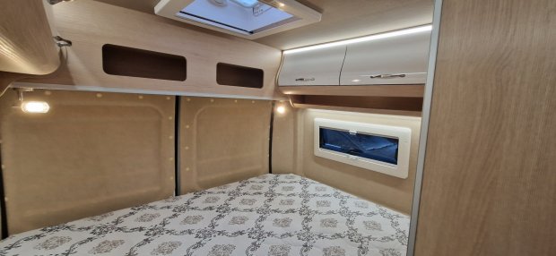 Kompaktowa sypialnia kampera: łóżko z wzorzystą pościelą, górne szafki i małe okno.