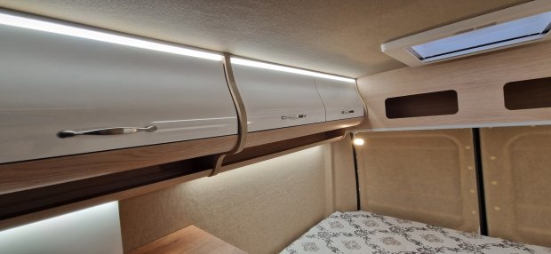 Białe połyskujące szafki z drewnym wykończeniem i oświetleniem LED nad łóżkiem kampera