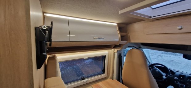 Wnętrze kampera: jasne drewniane szafki nad oknem, fotel kierowcy i kierownica.