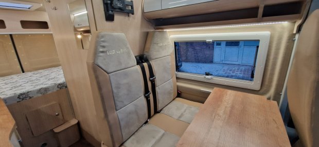 Wnętrze kampera: dwuosobowe tapicerowane siedzisko, stolik, okno i część łóżka.