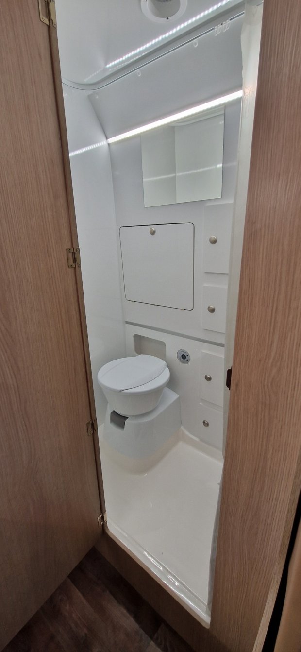 Mała biała łazienka w kamperze z toaletą, lustrem i drewnianymi drzwiami
