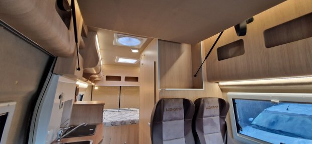 Wnętrze kampera z jasnym drewnem, aneksem kuchennym, łóżkiem i dwoma fotelami.