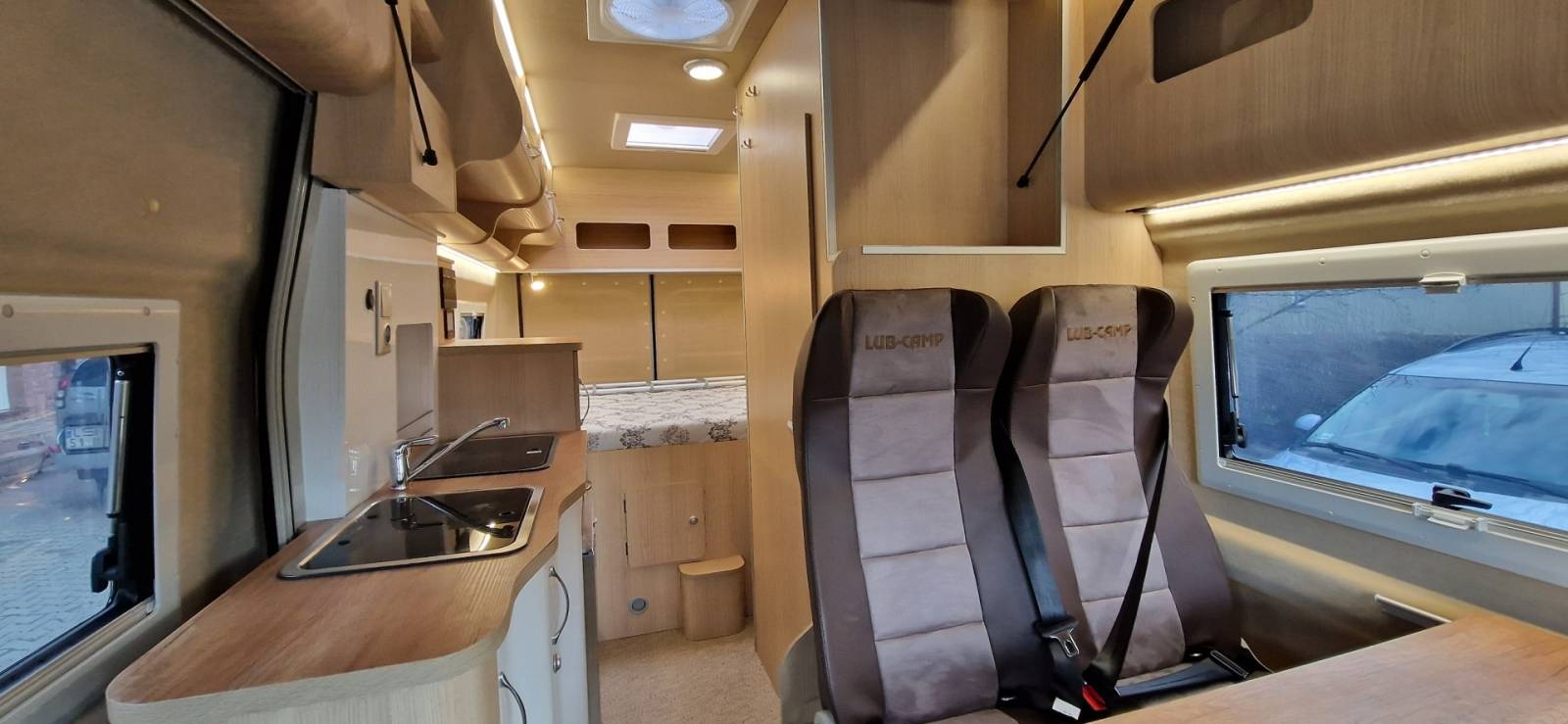 Wnętrze kampera: dwa fotele, aneks kuchenny i łóżko z tyłu, jasne drewno