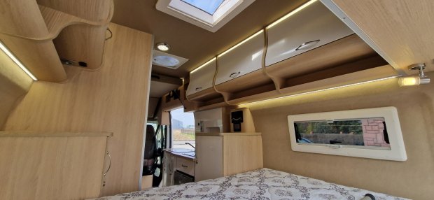 Wnętrze kampera z łóżkiem, drewnianymi szafkami, oknem i kuchenką.