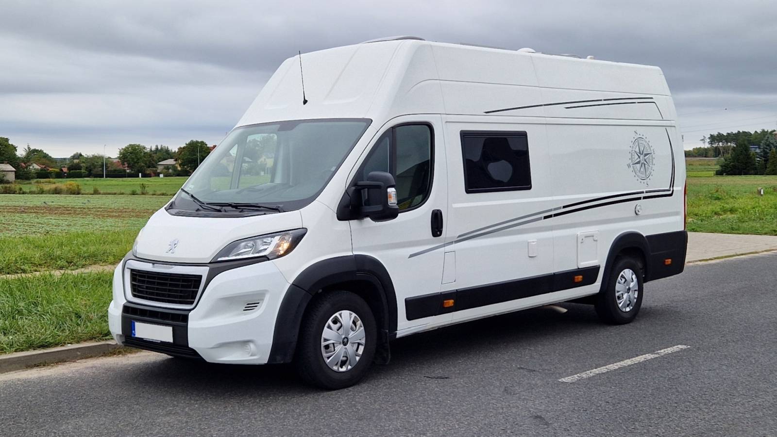 Biały kamper z oknem i kompasem na burcie zaparkowany przy drodze na tle pól