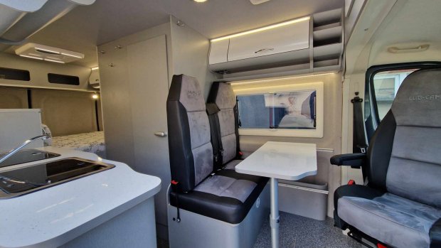 Wnętrze kampera z aneksem kuchennym, białym stolikiem i trzema tapicerowanymi siedzeniami