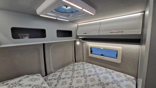 Wnętrze kampera: łóżko z wzorzystą pościelą, górne szafki, okno i świetlik.