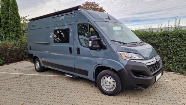 Szary kamperbus zaparkowany obok żywopłotu na kostce brukowej.