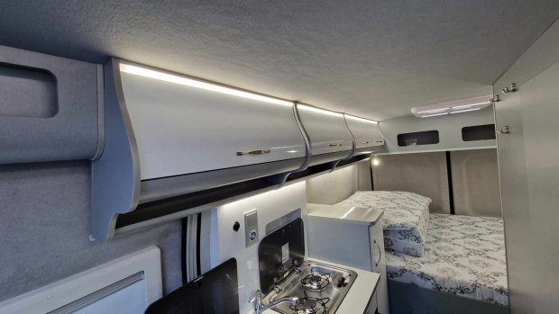 Wnętrze kampera z jasnymi szafkami, aneksem kuchennym i łóżkiem