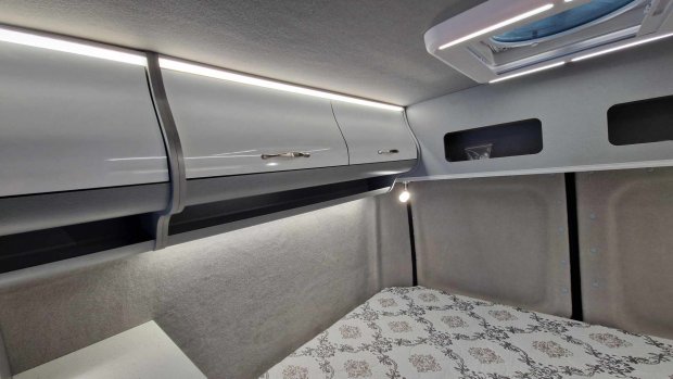 Wnętrze kampera: szare szafki sufitowe z oświetleniem, lampka i materac.