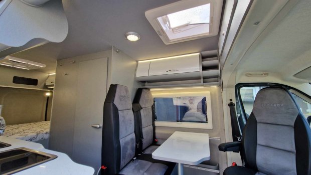 Wnętrze kampera z stołem, dwoma siedzeniami, fotelem kierowcy i górnymi schowkami.