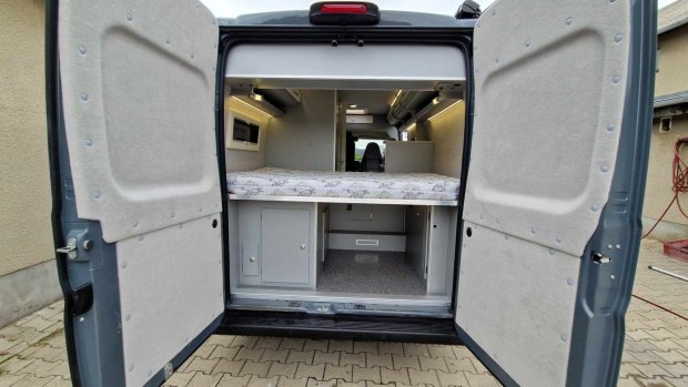 Tył kampera z otwartymi drzwiami, łóżkiem nad schowkami i szafkami