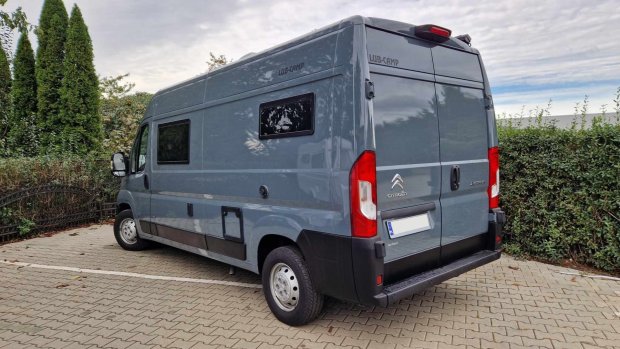 Szary kamper Citroën Jumper zaparkowany na kostce, widok z tyłu i boku, żywopłot w tle