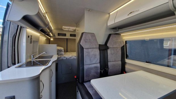 Wnętrze kampervanu: aneks kuchenny, stół, dwa fotele i łóżko w tle